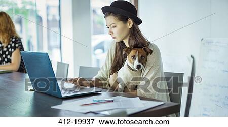 Stock Photo - Woman holding dog and working in office. Fotosearch