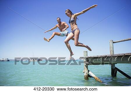 Stock Image - Couple jumping off wooden dock into water. Fotosearch