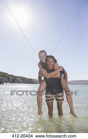 Picture - Woman being carried piggy back by boyfriend in waves . Fotosearch