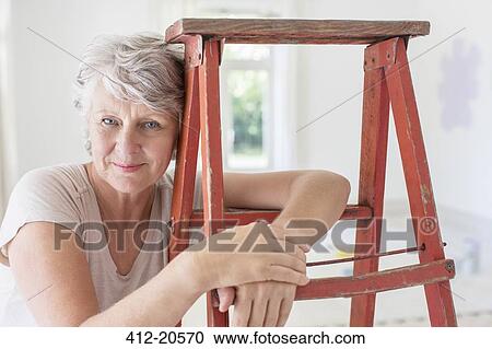 Stock Image - Older woman leaning on ladder in living space. Fotosearch