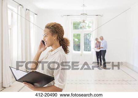 Stock Image - Woman talking on cell phone while couple looks at living room. Fotosearch