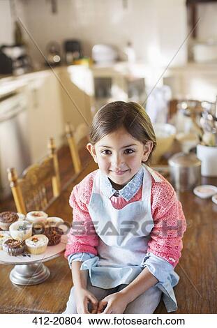 Picture - Young girl sitting on kitchen table near cupcakes . Fotosearch