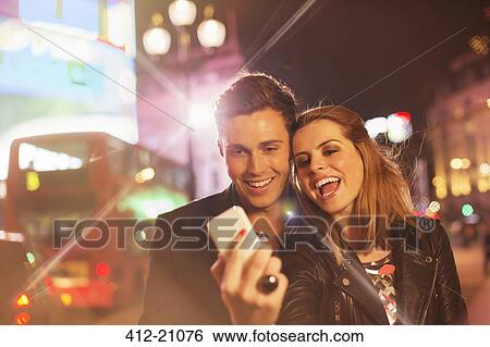 Couple taking cell phone picture together on city street at night View Large Photo Image Stock Photograph - Couple taking cell phone picture together on city street at night. Fotosearch