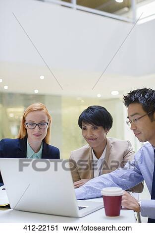 Stock Image - Business people gathered around laptop in office building cafe. Fotosearch