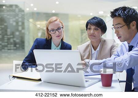 Stock Image - Business people gathered around laptop in office building cafe. Fotosearch