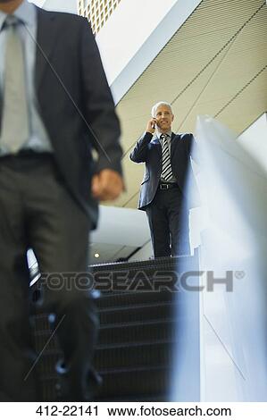 Stock Image - Businessman talking on cell phone at top of escalator in office. Fotosearch
