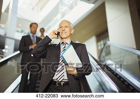 Stock Image - Businessman talking on cell phone going down escalator . Fotosearch