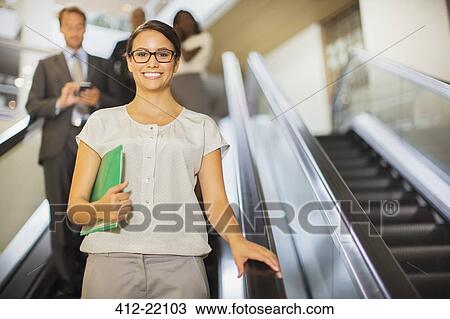 Stock Image - Businesswoman going down escalator in office building. Fotosearch