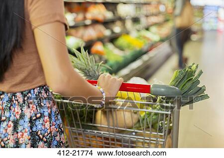 Picture - Close up of woman pushing full shopping cart in grocery store. Fotosearch