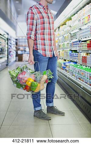 Stock Image - Man carrying full shopping basket in grocery store. Fotosearch