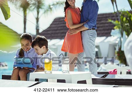 Brother and sister using digital tablet by resort swimming pool, parents embracing in background View Large Photo Image Stock Image - Brother and sister using digital tablet by resort swimming pool, parents embracing in background. Fotosearch