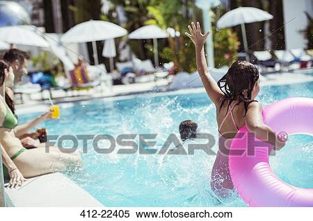 Stock Photography - Girl with pink inflatable ring jumping into swimming pool, family in background. Fotosearch