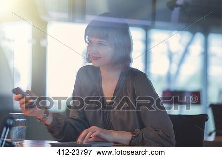 Stock Photo - Businesswoman holding cell phone at office desk. Fotosearch
