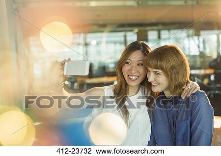 Stock Image - Businesswomen taking cell phone selfie in office. Fotosearch