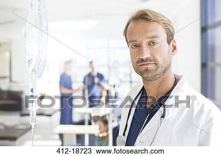 Stock Image - Portrait of mid adult doctor with colleagues in background, standing in hospital ward. Fotosearch