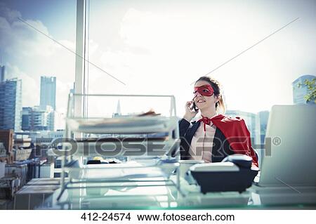 Picture - Businesswoman in cape and mask talking on phone at office desk. Fotosearch