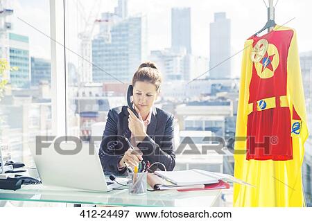 Stock Photo - Businesswoman working with superhero costume hanging in office. Fotosearch