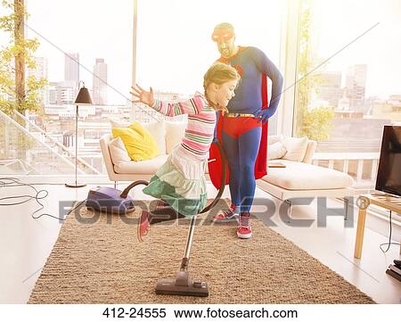 Stock Photography - Superhero father vacuuming while daughter jumps in living room. Fotosearch