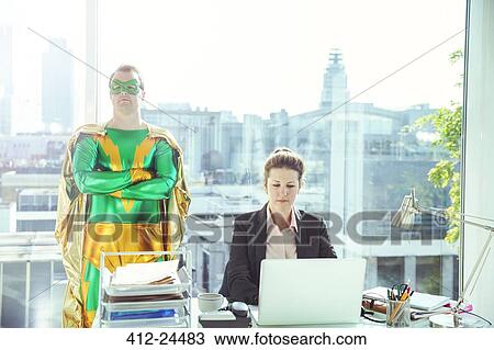 Stock Image - Superhero standing near businesswoman working in office. Fotosearch