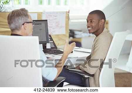 Stock Photo - "Men sitting at desk in office, talking and smiling". Fotosearch
