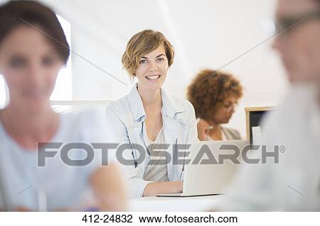Stock Image - "Portrait of woman sitting at desk, using laptop and smiling in office, colleagues in background". Fotosearch