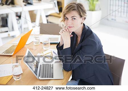 Portrait of woman sitting at desk with laptop in office View Large Photo Image Stock Photo - Portrait of woman sitting at desk with laptop in office. Fotosearch