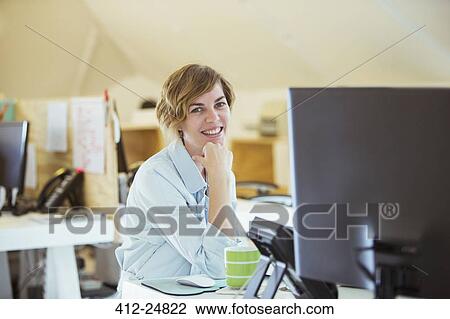 "Portrait of woman smiling in office, sitting at desk with computer" View Large Photo Image Stock Image - "Portrait of woman smiling in office, sitting at desk with computer". Fotosearch