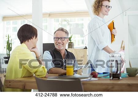 Stock Photo - Smiling office workers talking at desk during break. Fotosearch