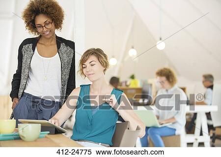 Stock Photography - Two office workers talking at desk using tablet. Fotosearch