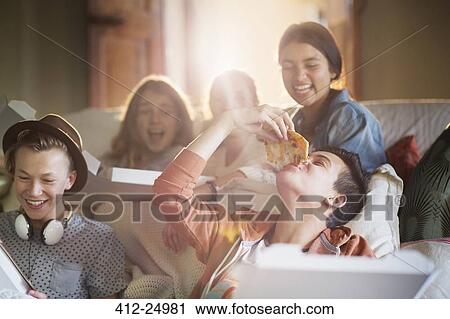 Stock Image - Group of teenagers eating pizza on sofa in living room. Fotosearch