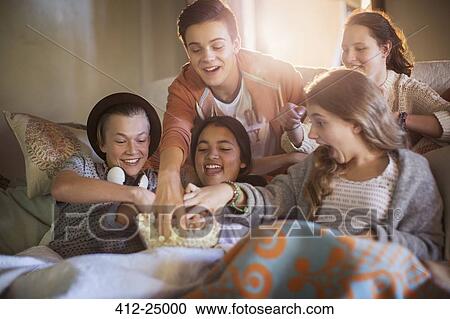 Stock Image - Group of teenagers eating popcorn on sofa in living room. Fotosearch