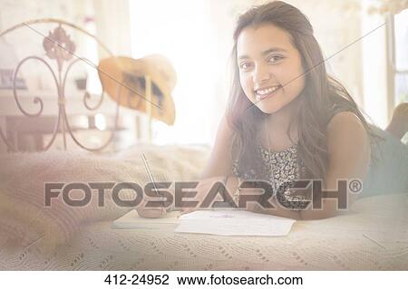 Stock Image - Portrait of smiling teenage girl lying on bed in bedroom. Fotosearch