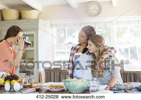 Stock Photo - Teenage girls photographing themselves in dining room. Fotosearch