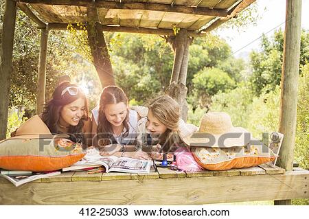 Stock Image - Three teenage girls reading magazine while lying in tree house in summer. Fotosearch