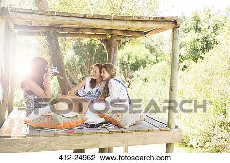 Stock Image - Three teenage girls taking photos in tree house in summer. Fotosearch
