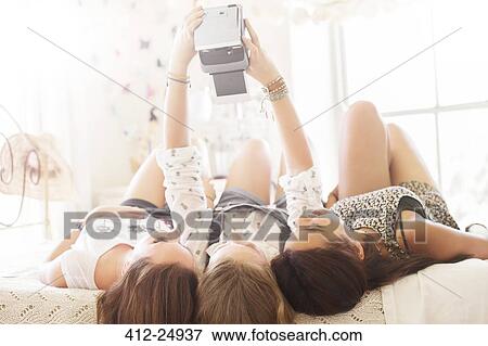 Stock Photo - Three teenage girls taking selfie while lying on bed in bedroom. Fotosearch