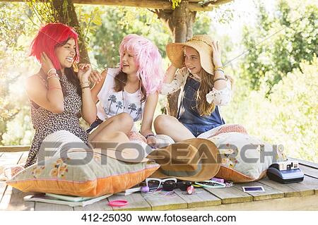 Stock Image - Three teenage girls wearing wigs and hat while sitting in tree house in summer. Fotosearch