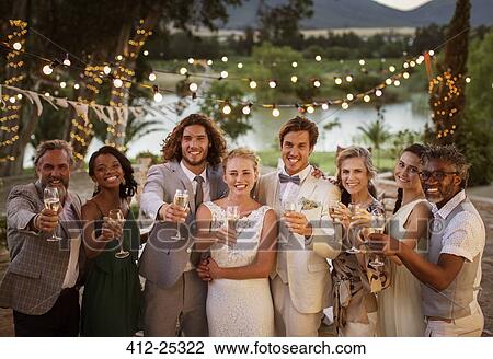 Stock Image - Portrait of young couple with guests toasting with champagne during wedding reception at dusk. Fotosearch