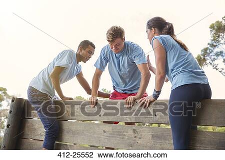 Stock Image - Teammates helping man over wall on boot camp obstacle course. Fotosearch