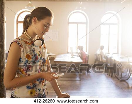 Stock Image - Casual businesswoman with headphones texting with cell phone in sunny office. Fotosearch