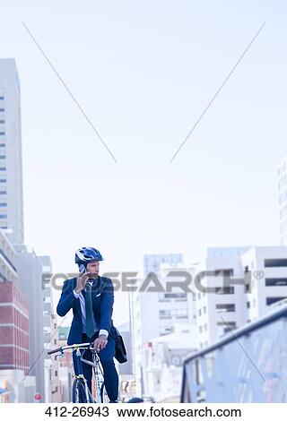 Stock Image - Businessman in suit and helmet talking on cell phone on bicycle in city. Fotosearch