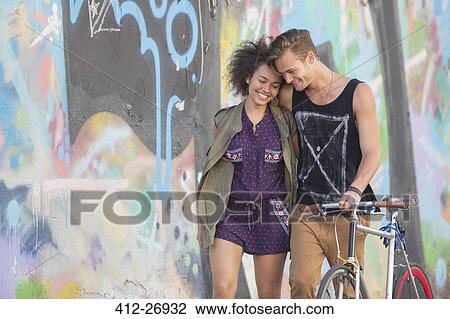 Stock Image - Couple hugging and walking with bicycle along urban graffiti wall. Fotosearch