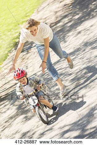 Mother pushing son with helmet on bicycle in sunny park View Large Photo Image Stock Photo - Mother pushing son with helmet on bicycle in sunny park. Fotosearch