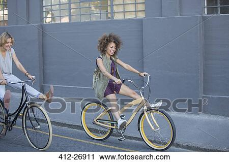 Stock Image - Playful women coasting on bicycles down urban street. Fotosearch