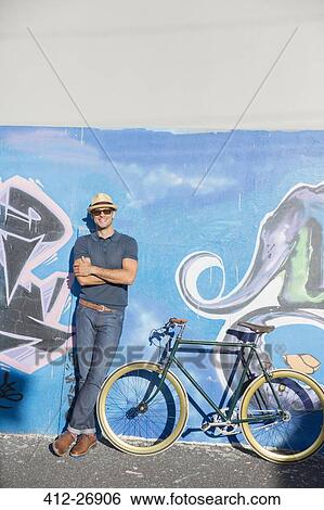 Portrait smiling man in hat and sunglasses leaning on urban graffiti wall next to bicycle View Large Photo Image Stock Photograph - Portrait smiling man in hat and sunglasses leaning on urban graffiti wall next to bicycle. Fotosearch