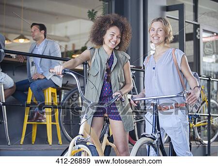 Stock Photo - Portrait smiling women on bicycles outside cafe. Fotosearch