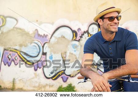 Stock Image - Smiling man in hat and sunglasses leaning on bicycle next to urban graffiti wall. Fotosearch