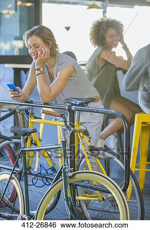Stock Photograph - Smiling woman leaning on railing texting with cell phone above bicycle. Fotosearch