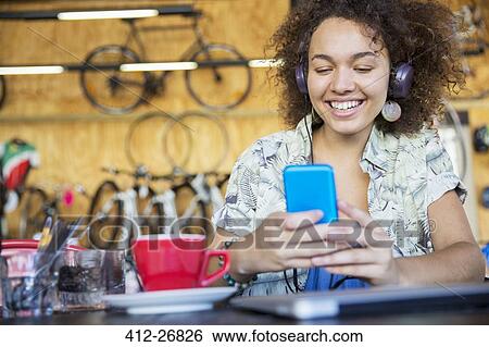 Stock Photograph - Smiling woman with headphones texting on cell phone in bike shop. Fotosearch