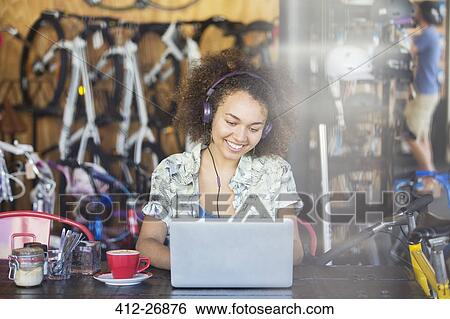 Stock Photograph - Smiling woman with headphones working at laptop in bicycle shop. Fotosearch